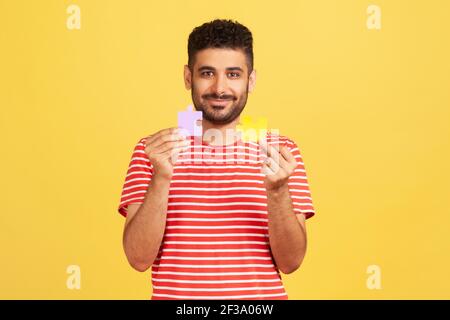 Uomo sorridente bearded in t-shirt a righe tenendo e mostrando due pezzi di puzzle, corrispondenza e puzzle di connessione, risolvere i problemi, creatività. Al coperto st Foto Stock