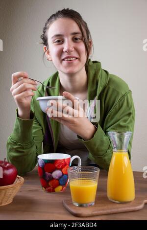 Giovane donna sorridente con una ciotola di cereali in mano, seduta, indossando abiti sportivi, dietro un tavolo da colazione con succo di frutta, mele e caffè, in po Foto Stock