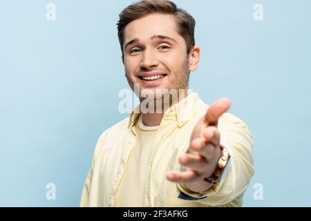 giovane allegro in camicia gialla con mano distesa sfocata isolato su blu Foto Stock