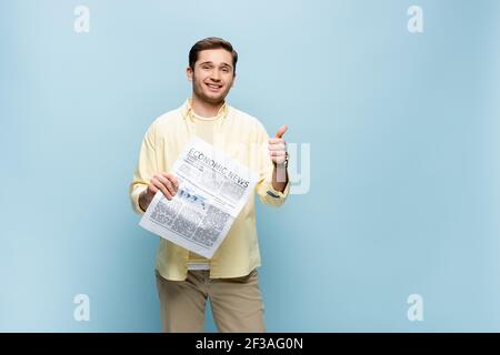 uomo sorridente in camicia che tiene il giornale e mostra il pollice in su isolato su blu Foto Stock