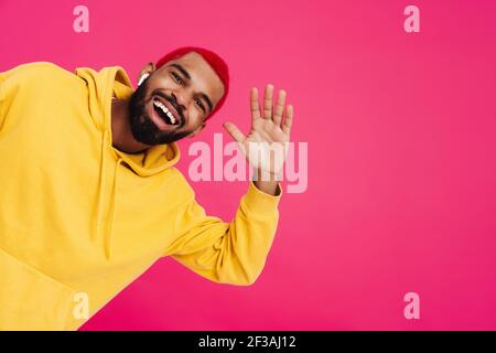 Felice funky giovane africano indossando una felpa con cappuccio ascoltando musica con auricolari wireless isolati su sfondo rosa, dicendo ciao Foto Stock