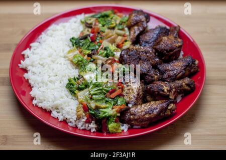 Un piatto di stile cinese a base di ali al forno servito con riso e verdure in una salsa densa e aromatica. Tutto servito su un piatto rosso adagiato su una w Foto Stock