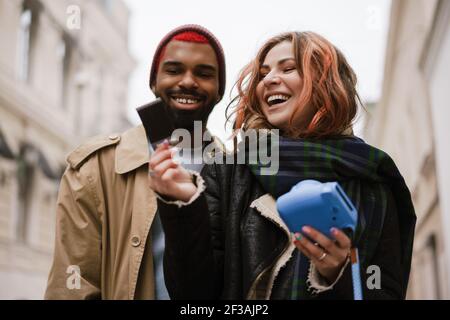 Una coppia gioiosa multinazionale che ride guardando una foto istantanea via della città Foto Stock