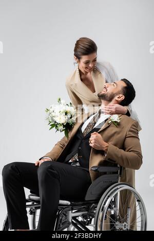 donna gioiosa con bouquet di nozze che abbraccia il collo di felice handicappato groom isolato su grigio Foto Stock