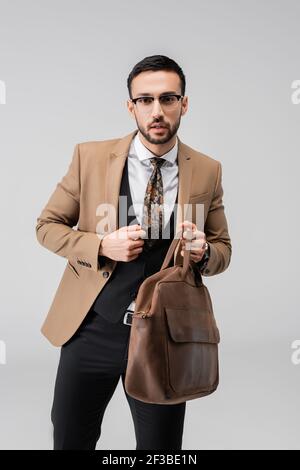 giovane uomo arabo in abiti eleganti che tengono la borsa in pelle isolata su grigio Foto Stock