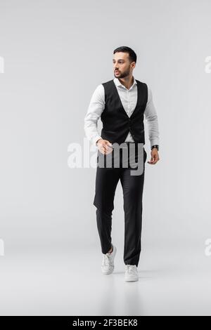 vista completa dell'uomo arabo alla moda in abiti eleganti e le sneakers che camminano sul grigio Foto Stock