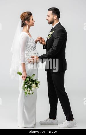 Vista laterale dello sposo musulmano che tiene la mano della sposa incinta su sfondo grigio Foto Stock