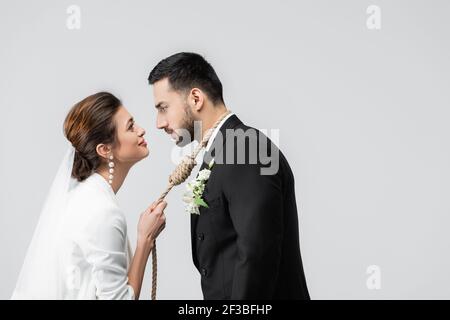 Vista laterale della sposa sorridente che tiene il nodo scivoloso sul collo di sposo musulmano isolato su grigio Foto Stock