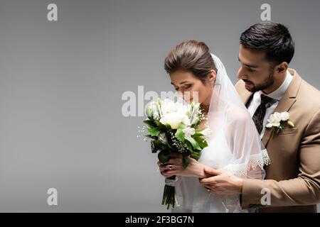 Sposa odore bouquet di nozze vicino allo sposo arabo isolato su grigio Foto Stock