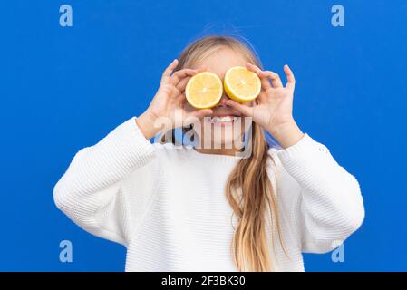 Ragazza con capelli biondi sopraffezionata in pullover bianco sorridente coprendo gli occhi con fette di limone luminose isolate su sfondo blu. Foto di alta qualità Foto Stock