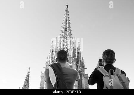 Due uomini, giovani e di mezza età, che ammirano e scattano foto con il telefono della torre gotica della Cattedrale di Barcellona dal suo tetto. Foto storica in bianco nero Foto Stock