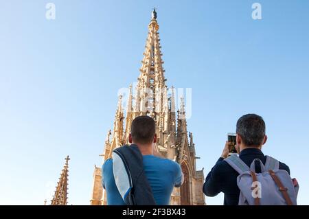 Due uomini (vista posteriore; irriconoscibile), giovani e di mezza età, ammirando e scattare foto con il telefono della torre gotica della Cattedrale di Barcellona dal suo tetto. Foto Stock