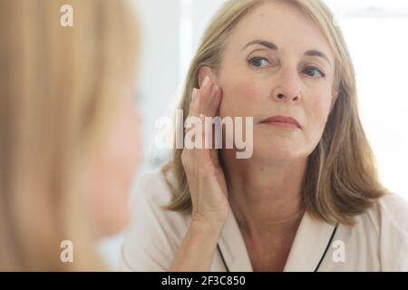 Donna caucasica anziana in piedi in bagno mettendo crema sul viso Foto Stock