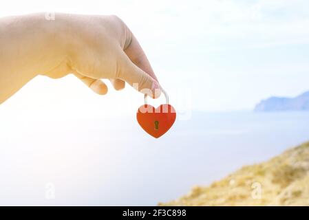Castello d'amore in rosso sullo sfondo del mare. La mano di un uomo tiene un piccolo lucchetto metallico. Relazione, amore e datazione concetto. Foto Stock