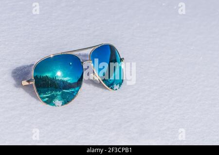 Occhiali da sole con lenti blu in cui il sole e il la foresta si riflette sulla neve Foto Stock