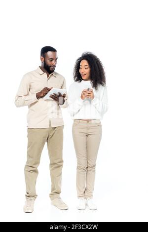 sorprende la donna afroamericana che tiene uno smartphone mentre guarda il digitale tavoletta in mani di marito su bianco Foto Stock