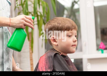 la mano femminile di un parrucchiere spruzzi acqua da un spray verde sulla testa di un bambino prima tagliare i capelli sullo sfondo di una finestra e. Foto Stock