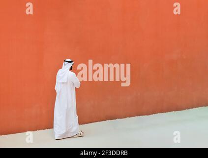 Uomo arabo con abiti tradizionali emirates che camminano all'aperto nel strada contro un muro arancione sfondo - etnia araba e. religione musulmana conc Foto Stock