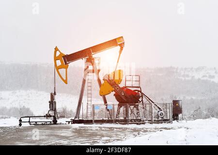 Pompa dell'olio. Attrezzature per l'industria petrolifera e del gas. Petrolio campo pompa martinetto e raffineria di petrolio in inverno con neve, montagne, foresta e tramonto in background Foto Stock