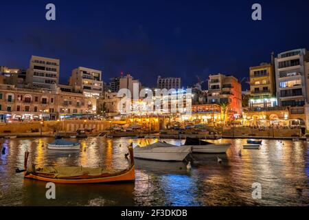 Paceville quartiere nella città di St Julian di notte a Malta, popolare luogo di vita notturna, barche nella baia di Spinola Foto Stock