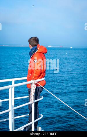 Uomo in abiti caldi in piedi sulla punta anteriore della barca, appoggiandosi sulle ringhiere e guardando via. Giorno di sole invernale sul mare. Foto Stock