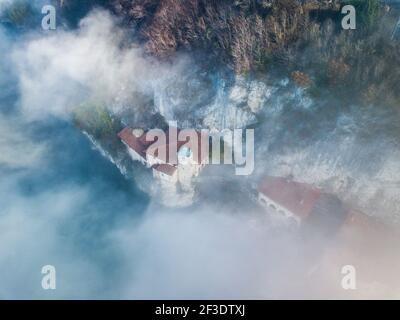 Vista frizzante dal drone. Edifici schiacciati tra la riva del lago e parete verticale di roccia. Sparse nebbia mattutina all'acqua. Foto Stock