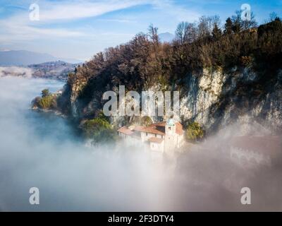 Vista aerea sugli edifici sotto la parete rocciosa dell'affioramento nel lago. Giorno di sole sopra la nebbia che si innalza dalla superficie dell'acqua. Foto Stock