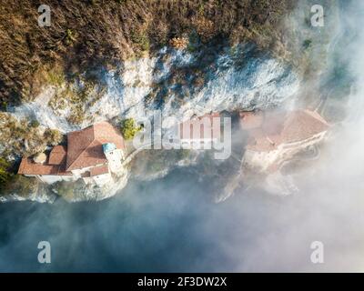 Vista dall'alto in basso su diverse case incastrate sulla roccia sopra la superficie dell'acqua. Il sole splende sui tetti sopra la nebbia sparsa. Foto Stock