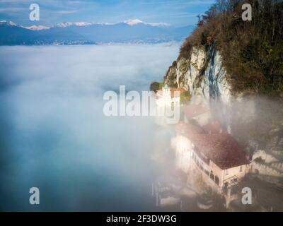 Vista aerea sulla valle allagata dalla nebbia. Alta cresta di montagna innevata in lontananza. Diverse case sotto parete di roccia che sporge sopra strato di nebbia. Foto Stock