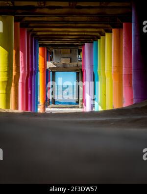 File di colonne colorate che sostengono il molo di legno sulla costa del mare. Vista dal soffietto della piattaforma. Arte di strada multicolore Foto Stock