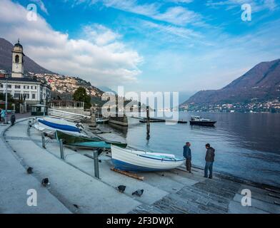 Due persone discutono accanto a barche a remi tirate a terra. Giornata fredda in città al lago. Cielo blu con nuvole bianche. Foto Stock