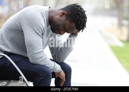 Profilo di un triste uomo nero che si lamenta da solo seduto una panchina in un parco Foto Stock