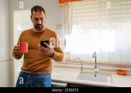 Bell'uomo bearded in un maglione arancione guardando lo smartphone in cucina mentre si ha una tazza di caffè. Concetto di tecnologia Foto Stock