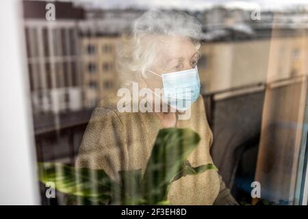 Donna anziana con maschera facciale che guarda fuori dalla finestra a. casa Foto Stock