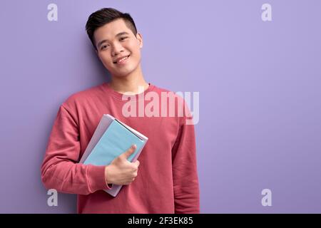 Ragazzo cinese con libro in mani godere di istruzione e l'università, in posa alla macchina fotografica, indossando camicia casual rossa, isolato su sfondo viola studio Foto Stock