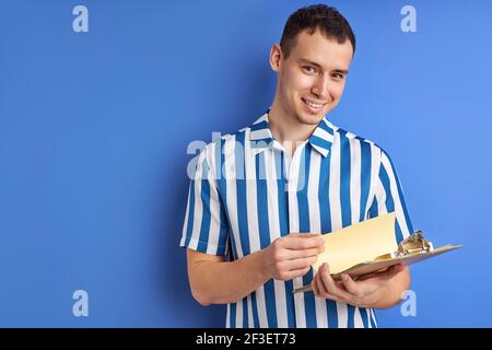 felice imprenditore o uomo d'affari con appunti isolati su sfondo blu, ritratto di bel ragazzo in camicia a righe blu guardando fotocamera avere Foto Stock