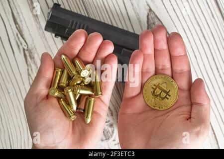 Pallottole e monete di colore giallo dorato in due mani, scrivania in legno offuscato con pistola nera sotto Foto Stock