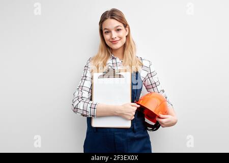 Donna ingegnere che tiene tablet di carta e casco, vestito in tute da costruttore uniforme e guardando fotocamera isolato su sfondo bianco studio. Contento Foto Stock