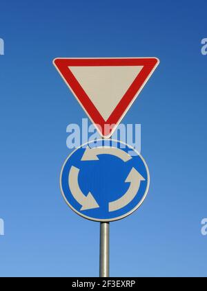 Un'immagine verticale di due segnali stradali sullo stesso Post - dare la strada e la rotonda blu indicazioni sulla sfondo blu cielo Foto Stock