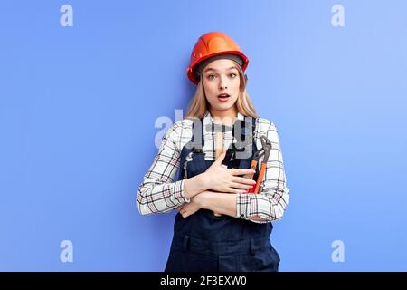 Ingegnere impegnato costruttore Donna guardando Camera wuth espressione sorpresa Tenere a mano strumenti regolabili isolati su uno studio blu indietro Foto Stock