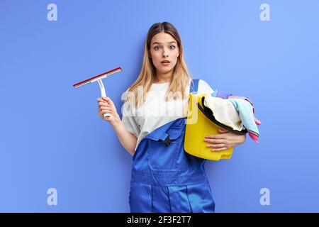 Donna che tiene il secchio con gruppo di materiali di pulizia. Giovane donna in tute in piedi in shock da sporcizia, attrezzature per la casa, primavera-pulizia, riassetto Foto Stock