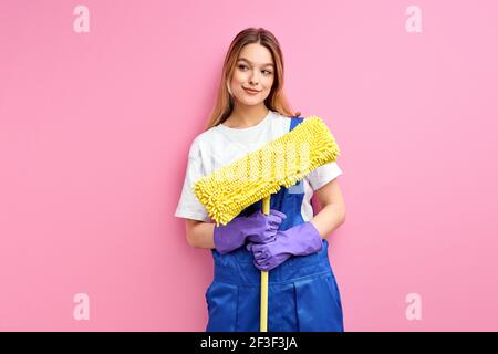 sorridente casa moglie di buon umore che tiene attrezzature per la pulizia, straccio per il pavimento, indossando tute blu uniforme isolato su sfondo rosa studio. portra Foto Stock