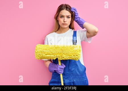 Donna più pulita indossare guanti tenendo mop su sfondo rosa isolato faccia seria pensare a domanda, idea molto confusa, tenendo testa. Portrai Foto Stock