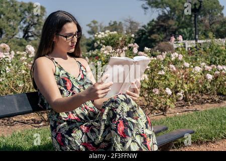 Bella giovane donna latina con bicchieri neri e abito fiorito in un parco concentrato a leggere un libro. Concetto di cultura. Foto Stock