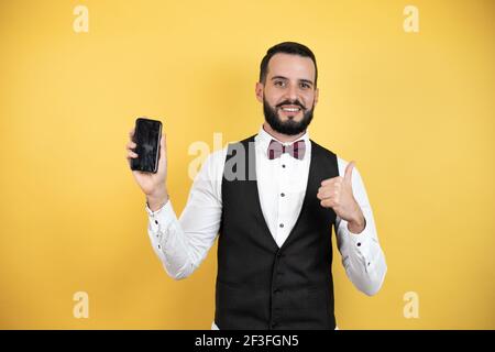 Giovane uomo con barba con cravatta ad arco e gilet sorridente felice di puntare con mano e dito sul lato sfondo giallo Foto Stock