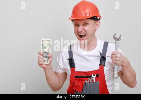 Lavoratore felice in uniforme rossa, casco protettivo che tiene il pacchetto di dollari, denaro contante e chiave su sfondo bianco. Lavoratore maschile per la pubblicità Foto Stock
