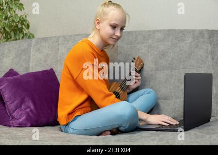 La ragazza adolescente impara a giocare l'ukulele e guarda nel suo laptop. Foto Stock