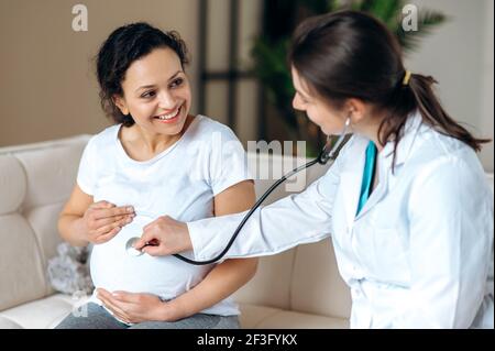 Salute di una donna incinta e di un bambino. La dottoressa effettua il controllo di felice gioiosa sorridente donna di razza mista incinta, ascoltando il battito cardiaco del bambino utilizzando stetoscopio, seduto sul divano in soggiorno Foto Stock