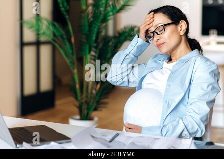 Esausta donna adulta in gravidanza mista seduta alla sua scrivania sente un mal di testa, è stressata, tiene la testa con la mano, gli occhi di chiusura, stanco di lavoro sedentario con il computer portatile, ha bisogno di una pausa e di riposo Foto Stock