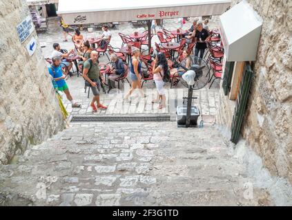 I turisti da una nave da crociera camminano oltre l'entrata a. La fortezza di Cattaro, guardando in su è scalinata di pietra antica come passano davanti a un caffè nella piazza Foto Stock
